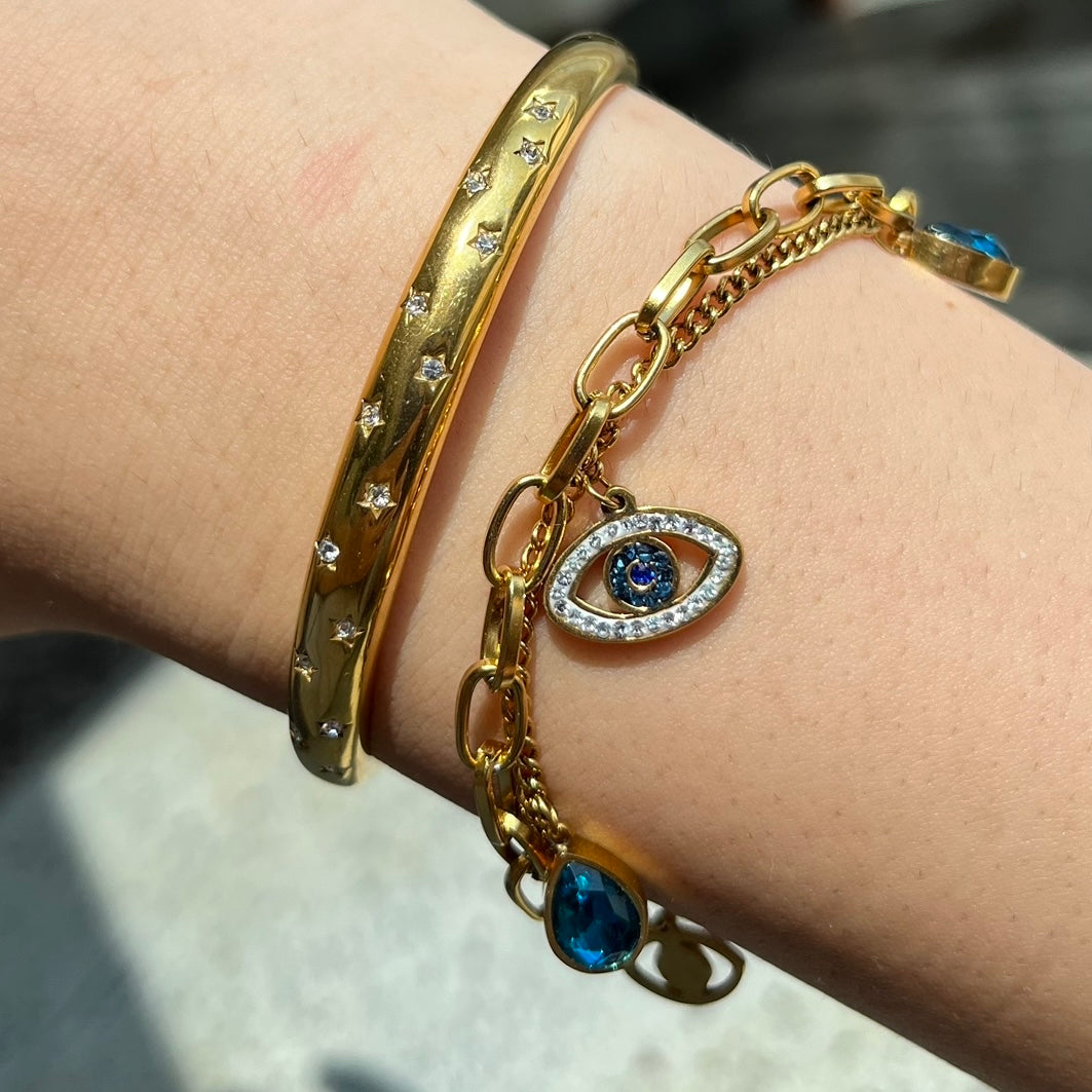 Eine Zusammensetzung von einem Armreif und einem Armband an einem Frauenarm. Der Armreif ist gold und hat eine abgerundete Form. In ihm sind Sterne eingraviert mit runden zirconia Steinen in der Mitte. Recht daneben ist ein Goldenes Armband mit einem Augenförmigen Anhänger und blauen zirkonia steinen.
