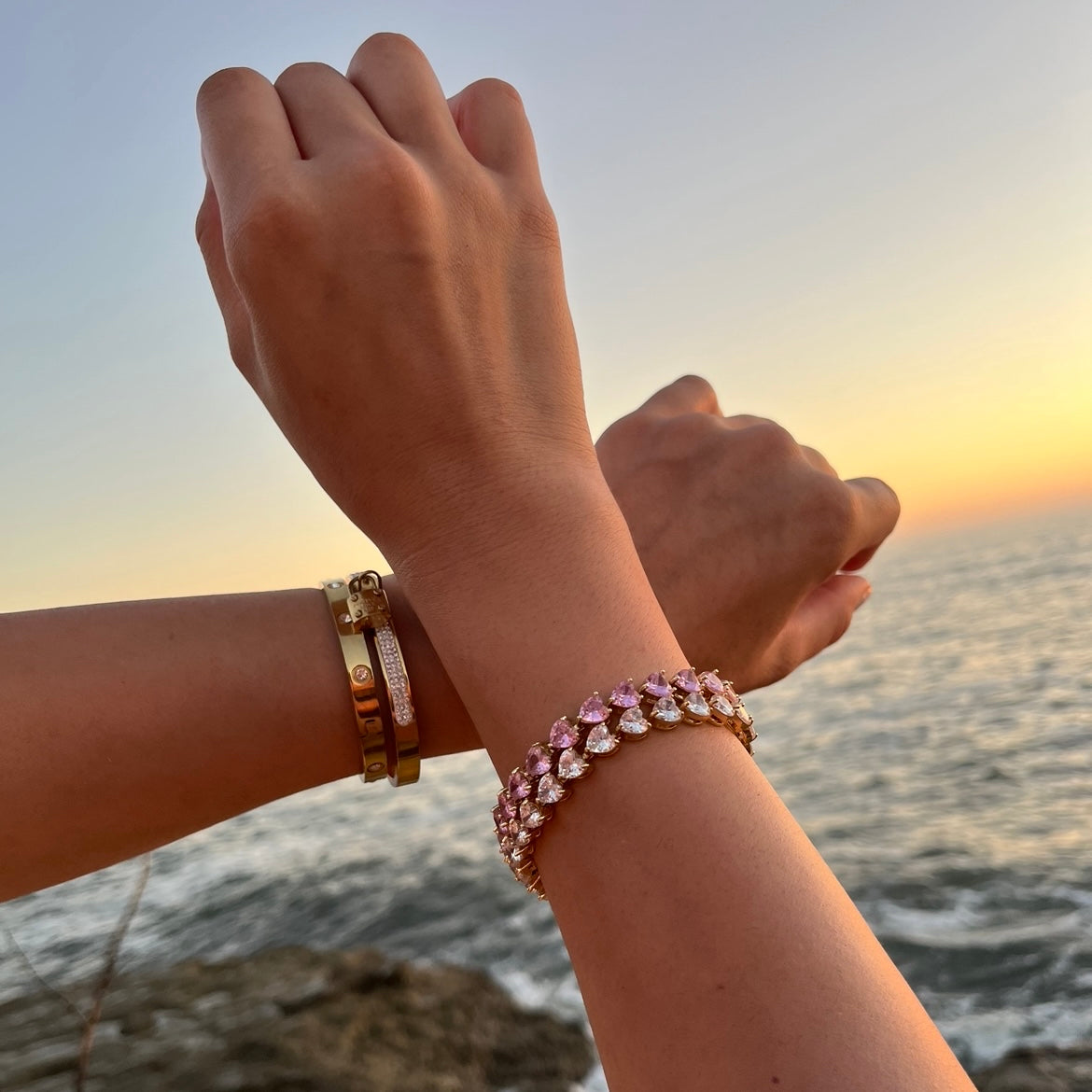 Goldenes Tennis Armband mit Rosa Zirkonia Herzen. Eingraviert mit dem Anna Carolina Logo. Das Armband hat Rosa herzen und ist ein Geschenk mit liebe für die Partnerin, Beste Freundin oder kann auch als Freundschaftsarmband getragen werden. Werden von zwei frauenarmen getragen. Im Hintergrund Meer und Abendsonne.