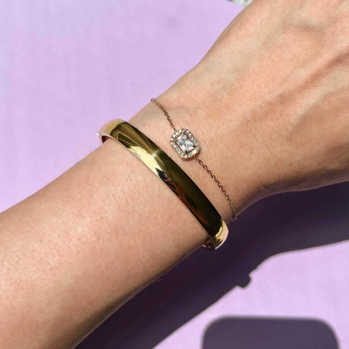 Breiter goldener Armreif in Hochglanz an einem Frauenarm, neben einem feinen Armband mit Rechteckigem Zirconia. Ein lila Hintergrund, passend zu der Marken Farbe von Anna Carolina