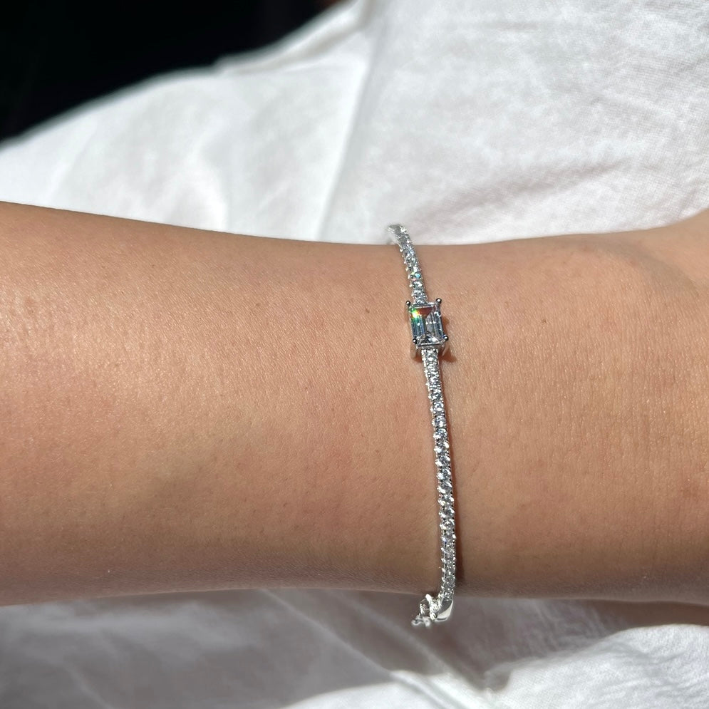 Schmaler feiner Armreif aus 925 Sterling Silber mit rhodium beschichtet um einen schmalen Frauenarm. Der Armreif ist mit unzähligen kleinen zirconia Steinchen besetzt die wunderschön schimmern. In der Mitte ist ein großer Zirconia Stein angebracht mit elegantem Smaragdenschliff. Der Armreif ist mit 925 eingraviert als symbol von Qualität. Der Armreif ist ein hochwertiges Schmuck Geschenk für Frauen die zeitlose Eleganz und Langlebigkeit Schätzen. Im Hintergrund ist ein weißer Rock zu sehen.