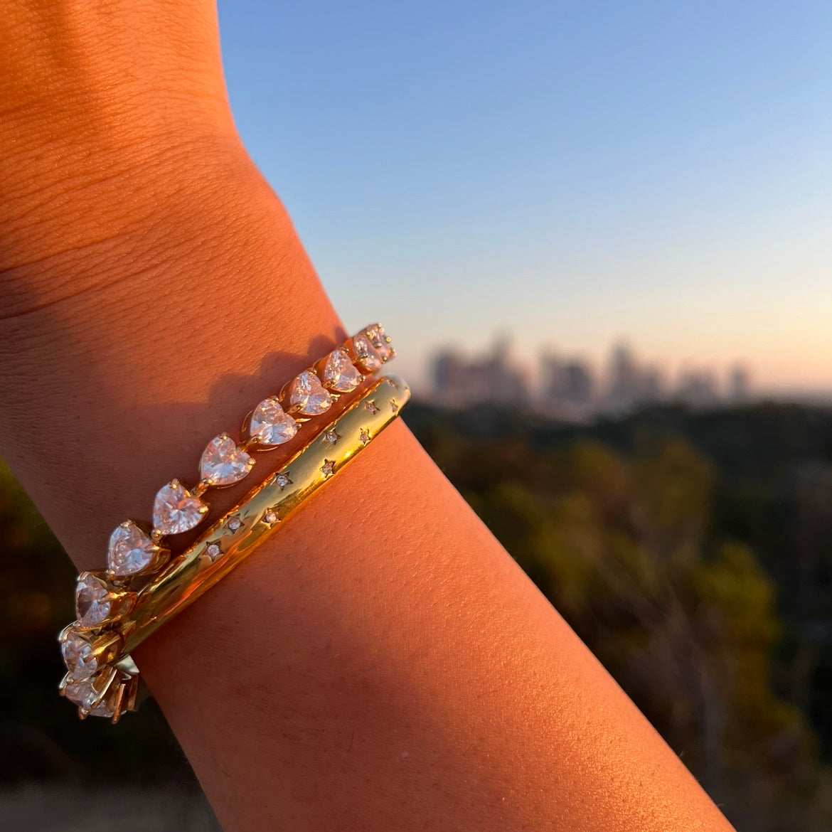 Ein Tennisarmband mit weißen Zirkonia  Herzen in Gold neben einem goldenen rundem Armreif mit weißen zirconia Sternen, an einem Arm einer Frau. Im Hintergrund ist die Skyline von Los Angeles zu sehen. Die zironia steine schimmern elegant in der rötlichen Abendsonne.