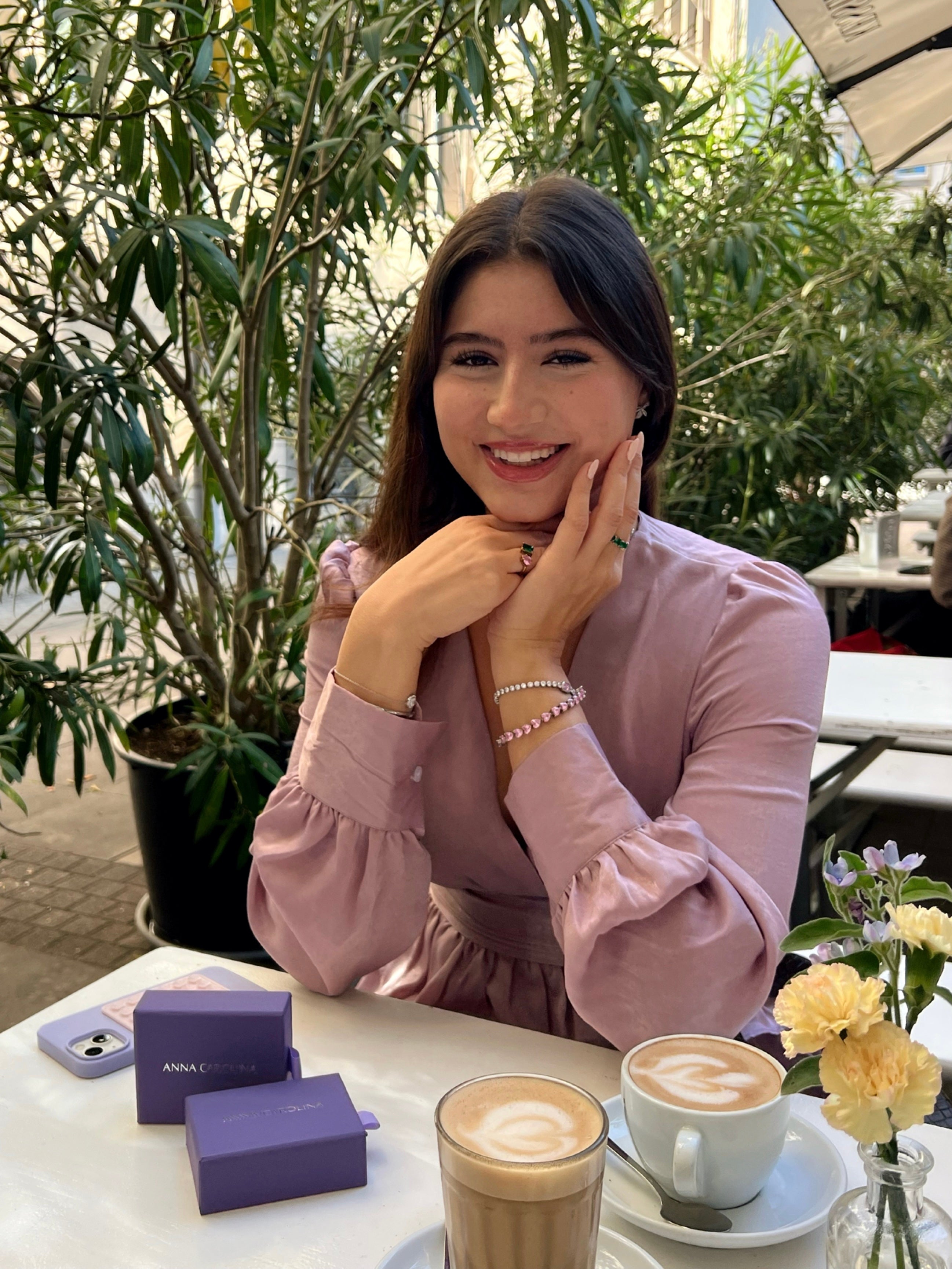 Smiling girl sitting in a cafe wearing a purple dress and a silver tennis bracelet with pink heart shaped stones. On the table are yellow and purple flowers, as well as two purple Anna Carolina jewellery boxes. On her hands she is wearing two gold rings with green zirconia stones.