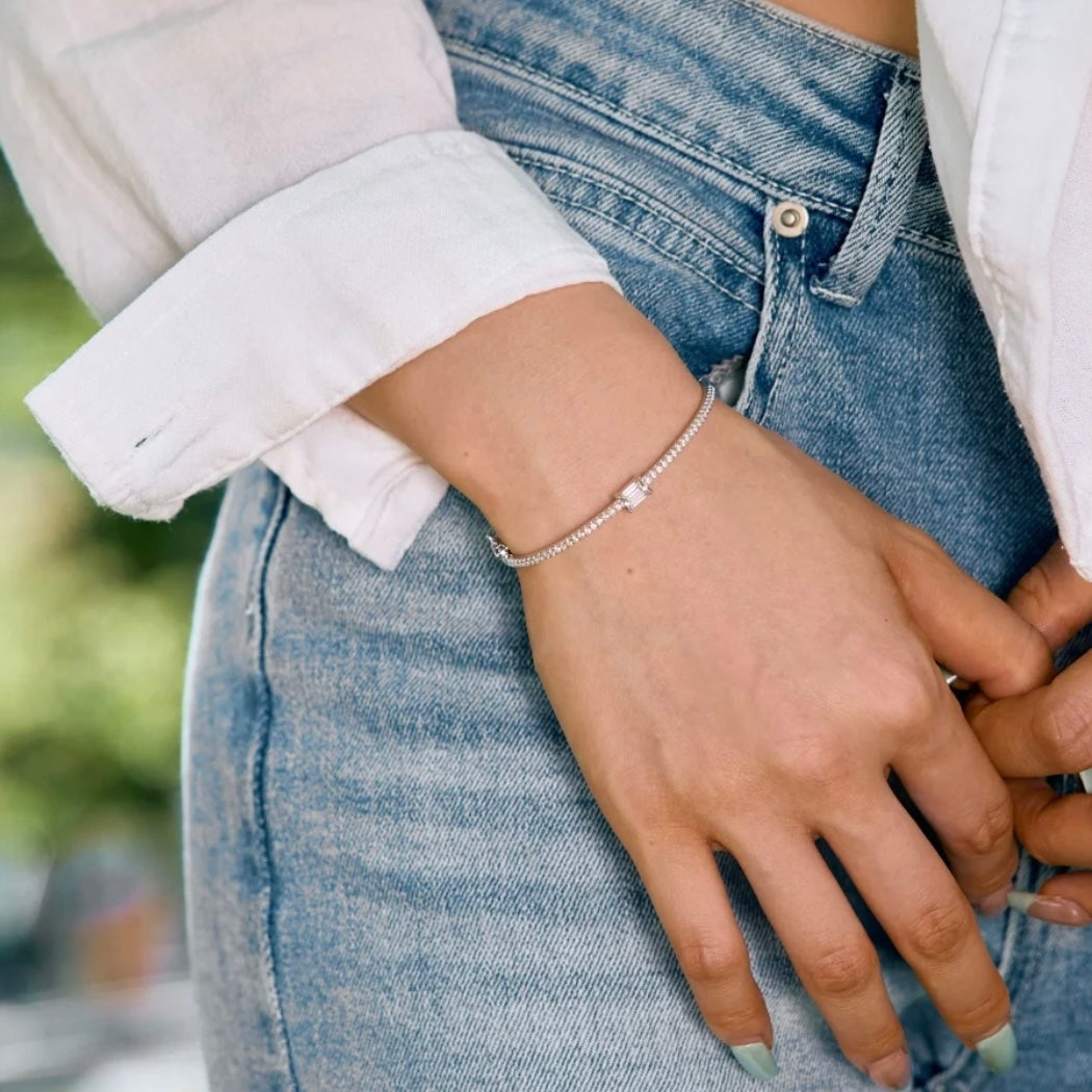 Schmaler feiner Armreif aus 925 Sterling Silber mit rhodium beschichtet. Der Armreif wird von einer elegant gekleideten Frau getragen.  Der Armreif ist mit unzähligen kleinen zirconia Steinchen besetzt die wunderschön schimmern. In der Mitte ist ein großer Zirconia Stein angebracht mit elegantem Smaragdenschliff. Der Armreif ist mit 925 eingraviert als symbol von Qualität. Der Armreif ist ein hochwertiges Schmuck Geschenk für Frauen die zeitlose Eleganz und Langlebigkeit Schätzen.  